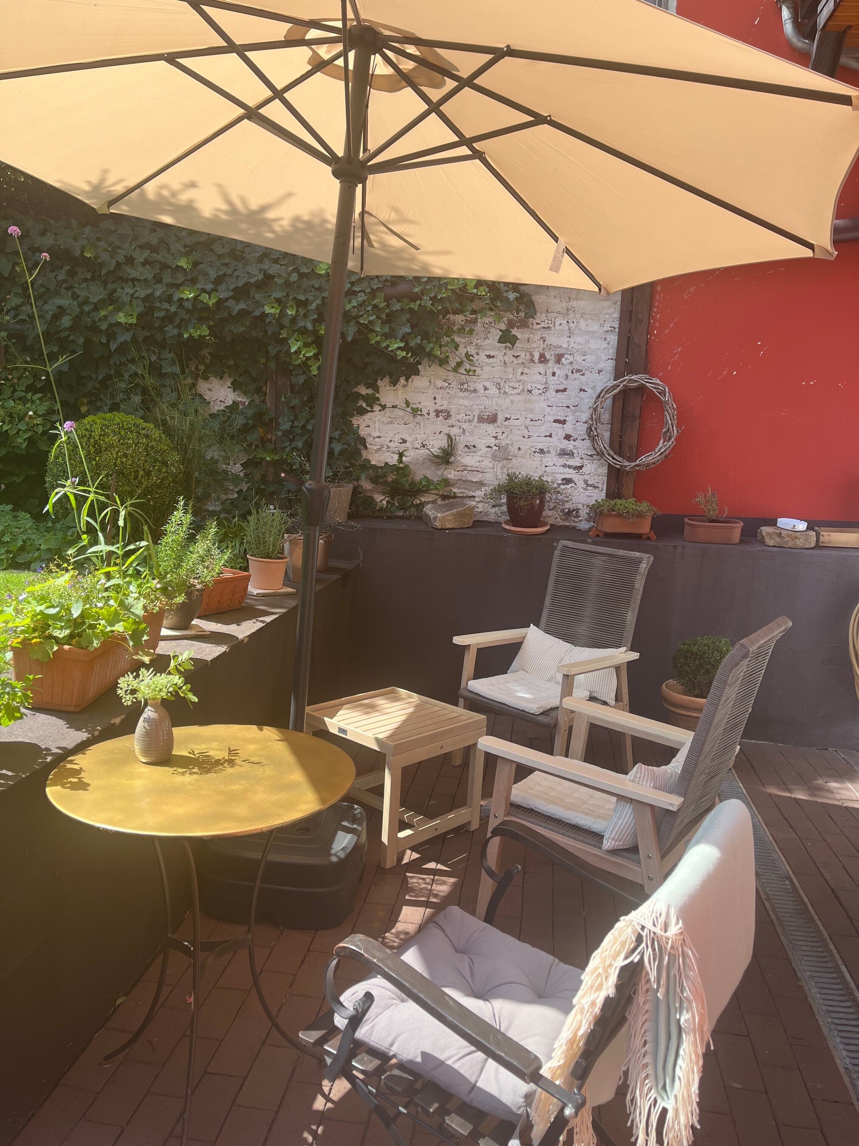 Una terrazza esterna con un tavolo rotondo, due sedie, un ombrellone grande e diverse piante in vaso.