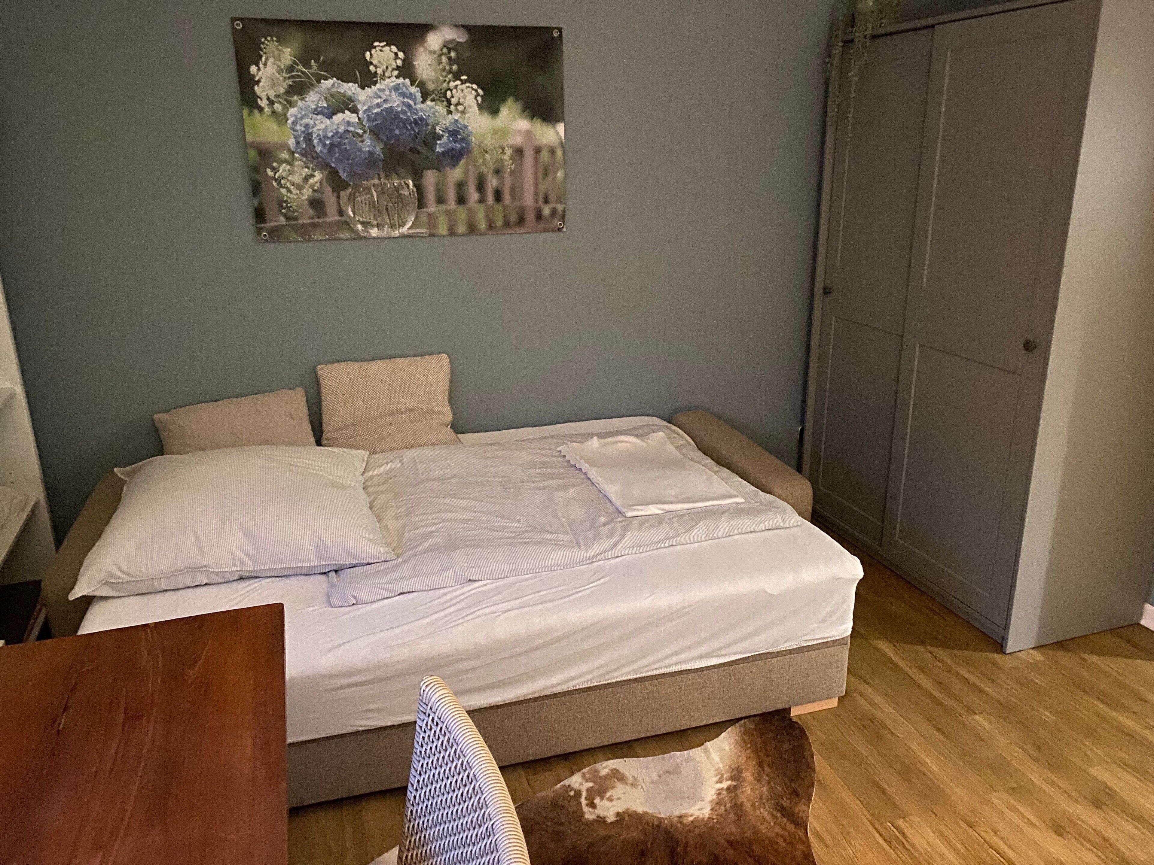 A bedroom with a bed, pillows, a wooden floor, a wardrobe, and a painting on the wall.