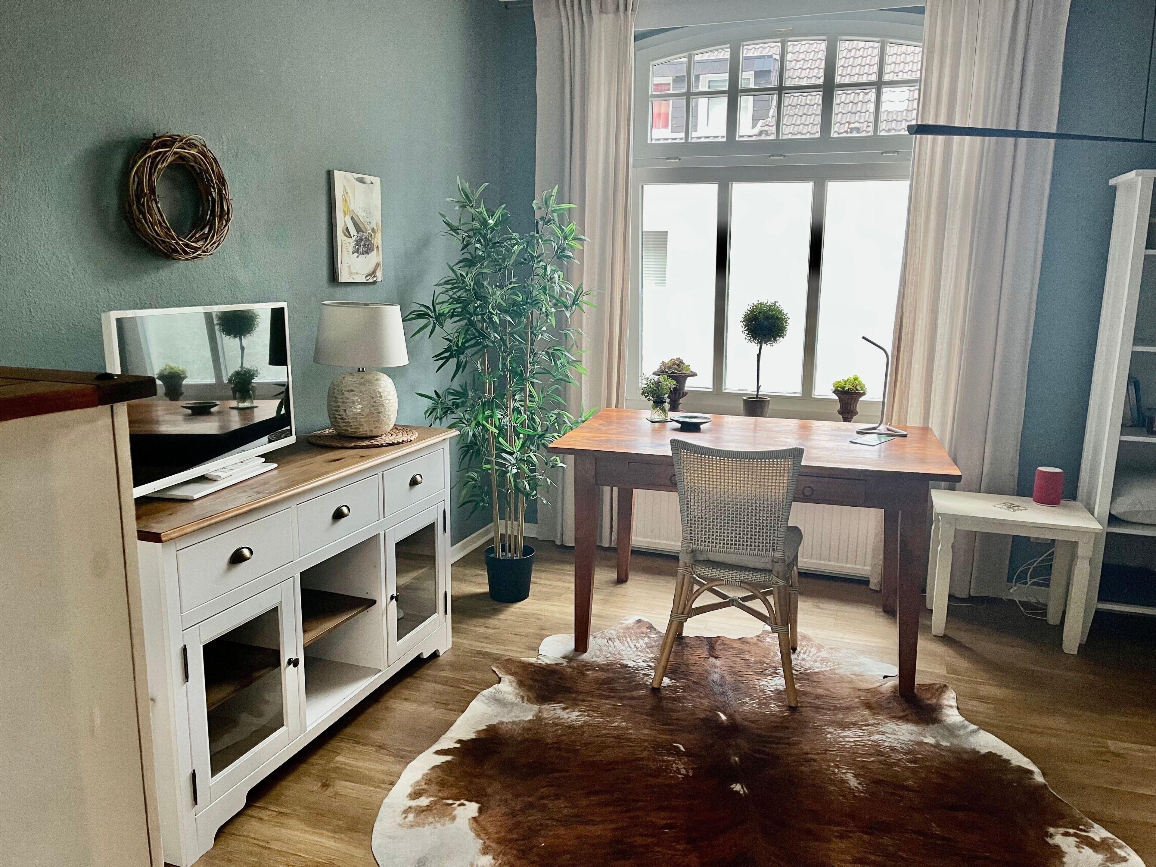 Ein Zimmer mit Schreibtisch aus Holz, einem Sessel, einem Fernseher, einer Lampe, einer Zimmerpflanze und einem Kuhfellteppich.