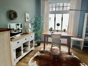 Ein Zimmer mit Schreibtisch aus Holz, einem Sessel, einem Fernseher, einer Lampe, einer Zimmerpflanze und einem Kuhfellteppich.