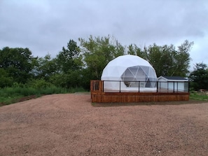 Une structure en forme de dôme géodésique, dotée d’une terrasse et d’une rambarde en bois, entourée de gravier et d’arbres.