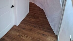 Un pasillo estrecho con suelo de madera, una puerta blanca y un ventanal con cortina blanca.