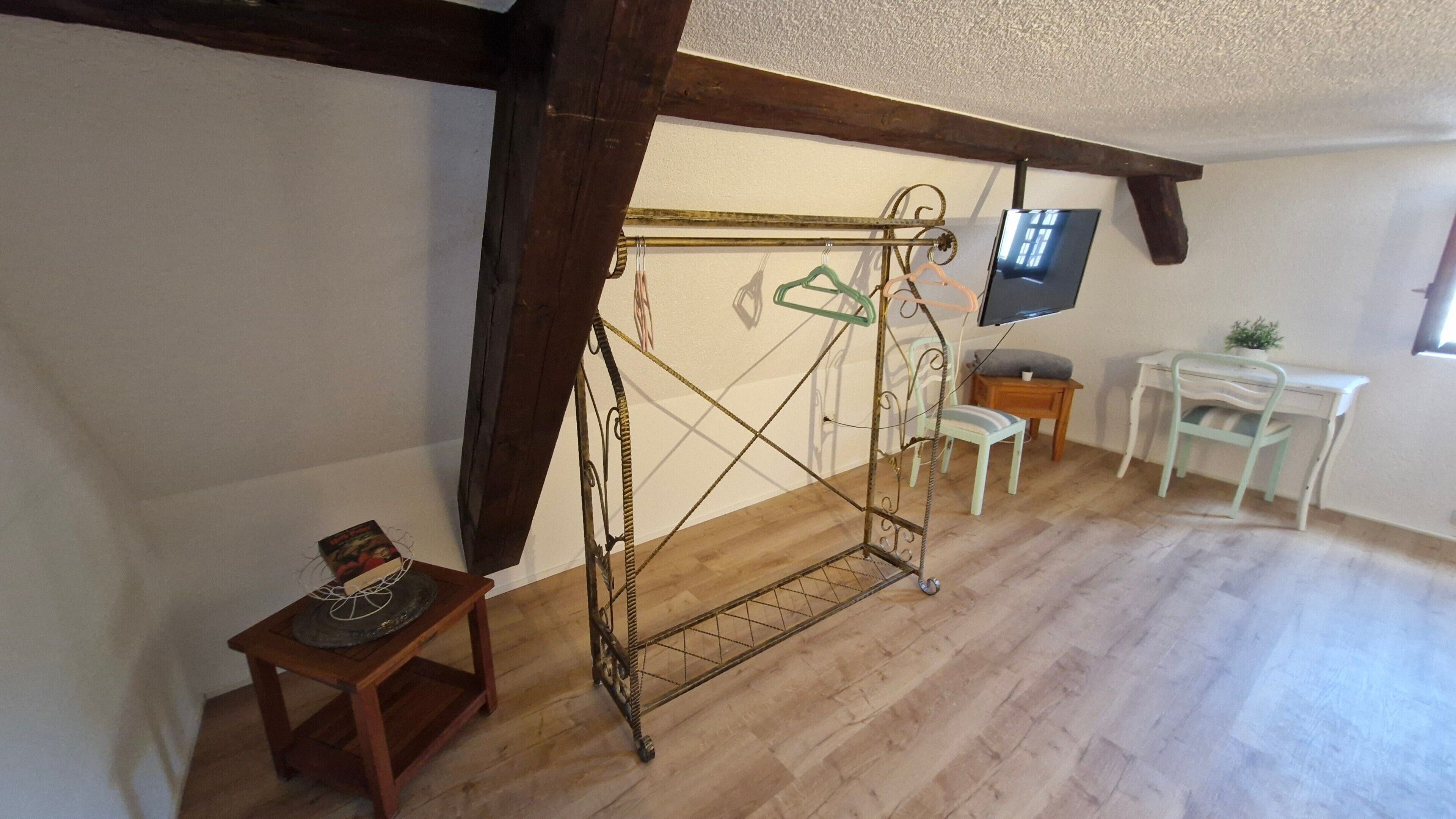 Une pièce avec un plancher en bois, un portant à vêtements en métal, une petite table en bois sur laquelle se trouve un livre, un bureau blanc avec une chaise et un téléviseur fixé au mur.