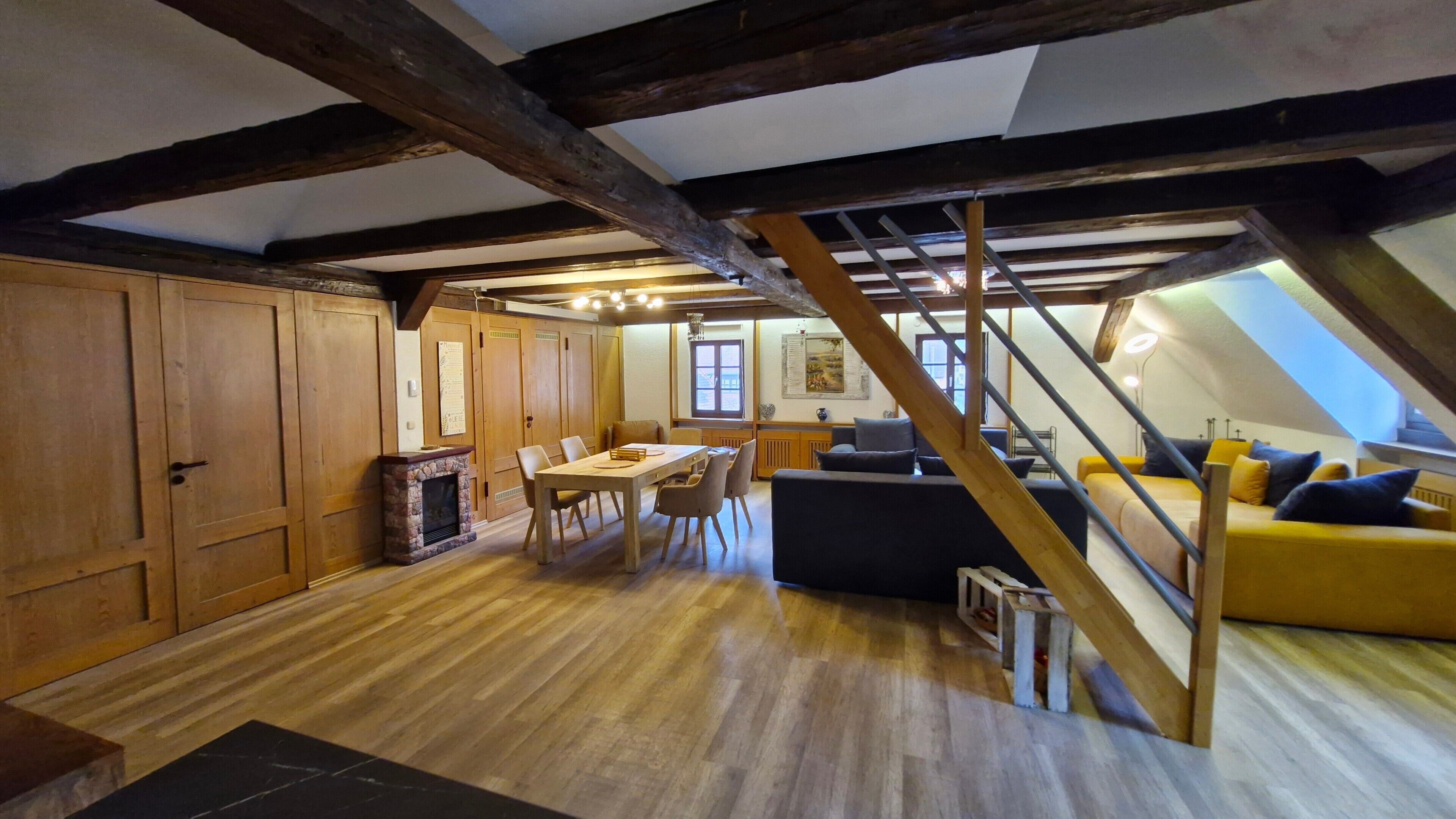 Une pièce de style « loft » avec des poutres en bois apparentes, un coin repas avec une table et des chaises, un canapé et un escalier.