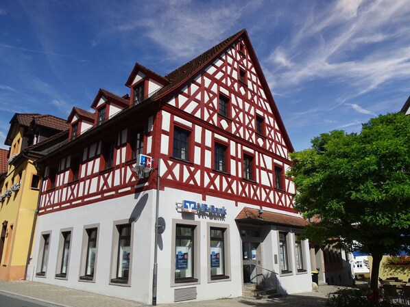 Ein traditionelles Fachwerkhäuschen mit sichtbarem Bankschild, Fenstern und einer Treppe zum Eingang.