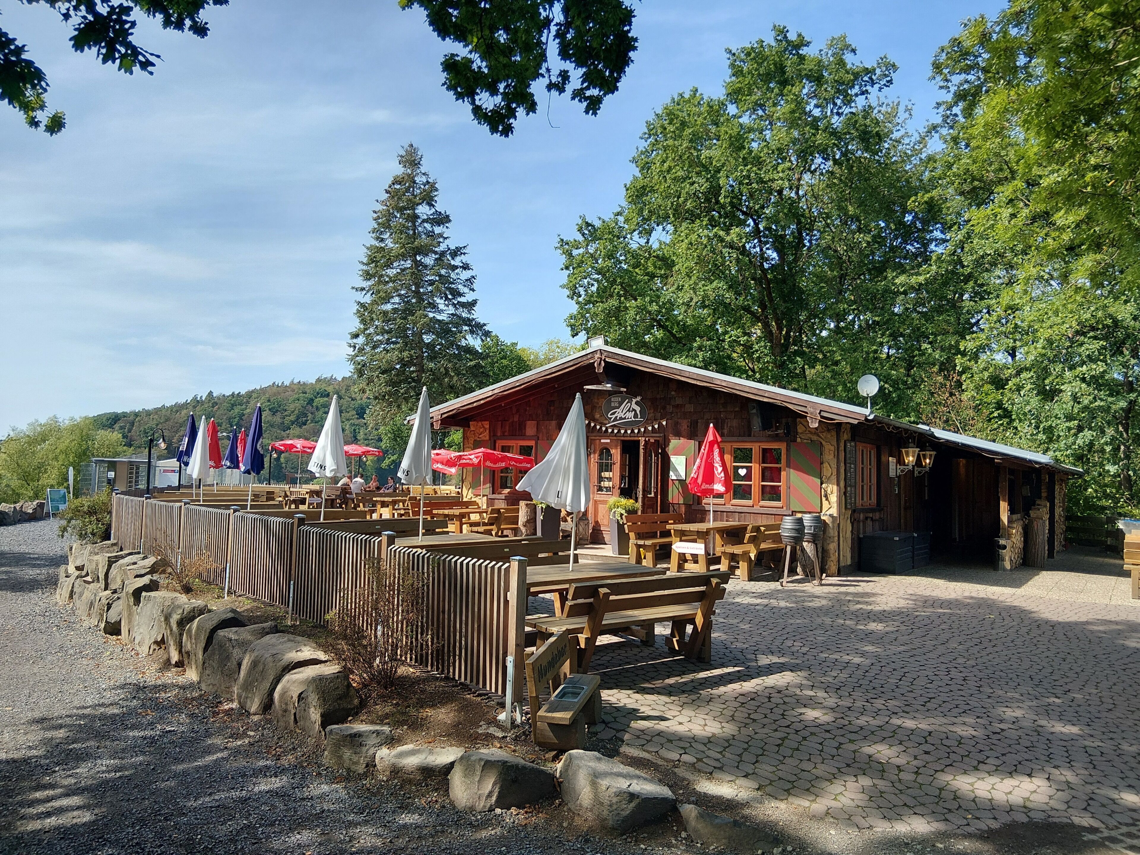 Ein rustikales Holzgebäude mit Außenplätzen und Sonnenschirmen, umgeben von Bäumen und einem Kiesweg.