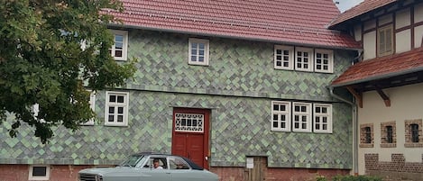 A two-story building with a red door and multiple windows, a cobblestone street, and a vintage car parked in front.