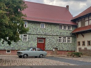 Ein zweistöckiges Gebäude mit roter Tür und mehreren Fenstern, eine gepflasterte Straße und ein klassisches Auto davor.