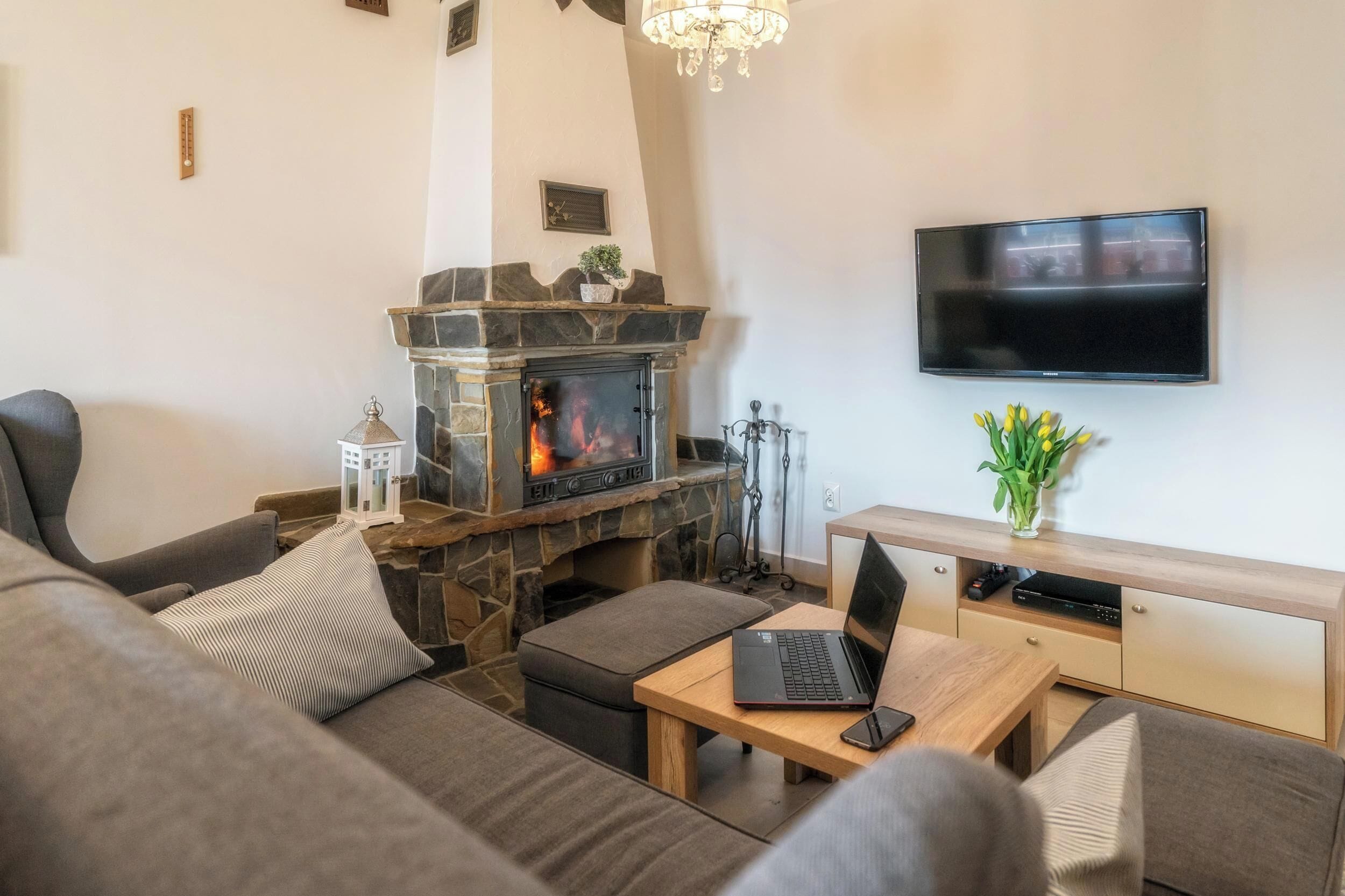 A living room with a stone fireplace, a flat-screen TV mounted on the wall, a laptop on a wooden table, and a sofa with cushions.