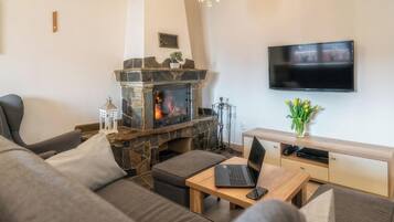 A living room with a stone fireplace, a flat-screen TV mounted on the wall, a laptop on a wooden table, and a sofa with cushions.
