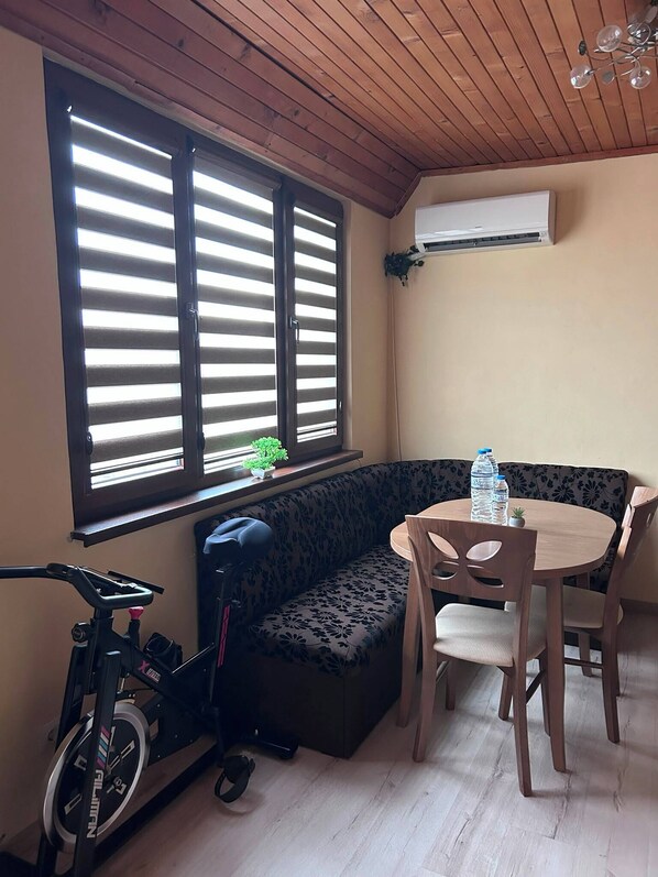 A room with a wooden ceiling, a window with blinds, a sofa, a table with chairs, and an air conditioning unit.