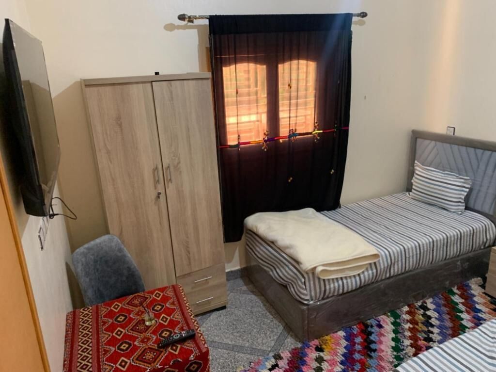 A bedroom with a bed, a wardrobe, a television, and a small table with a patterned tablecloth.