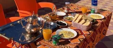 An outdoor dining area with a table set for a meal, surrounded by orange chairs, and a view of trees and buildings in the background.