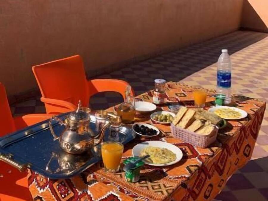 An outdoor dining area with a table set for a meal, surrounded by orange chairs, and a view of trees and buildings in the background.