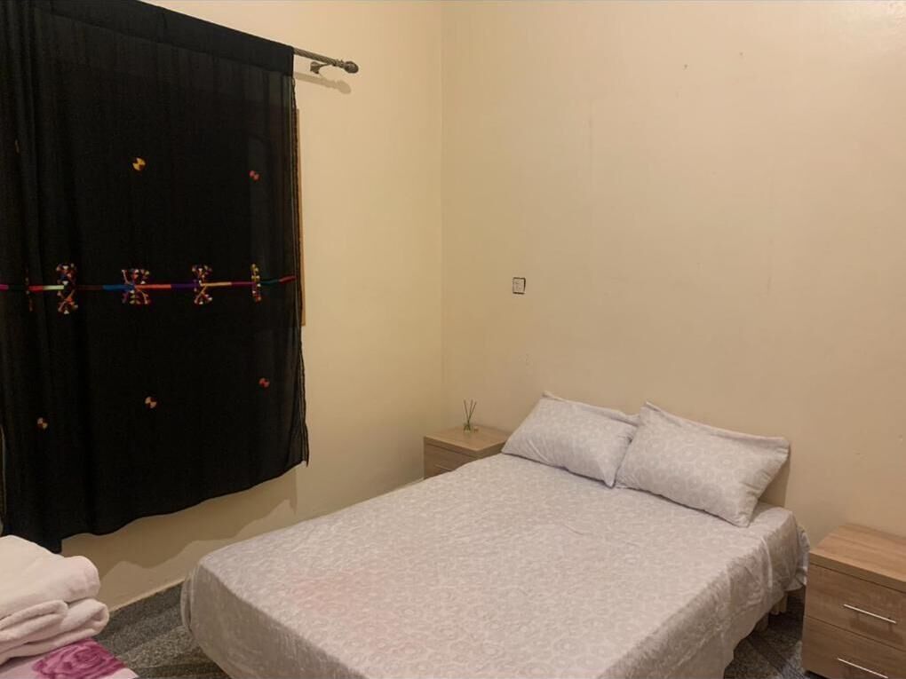 Een slaapkamer met een bed, nachtkastje, ladekast en gordijn.