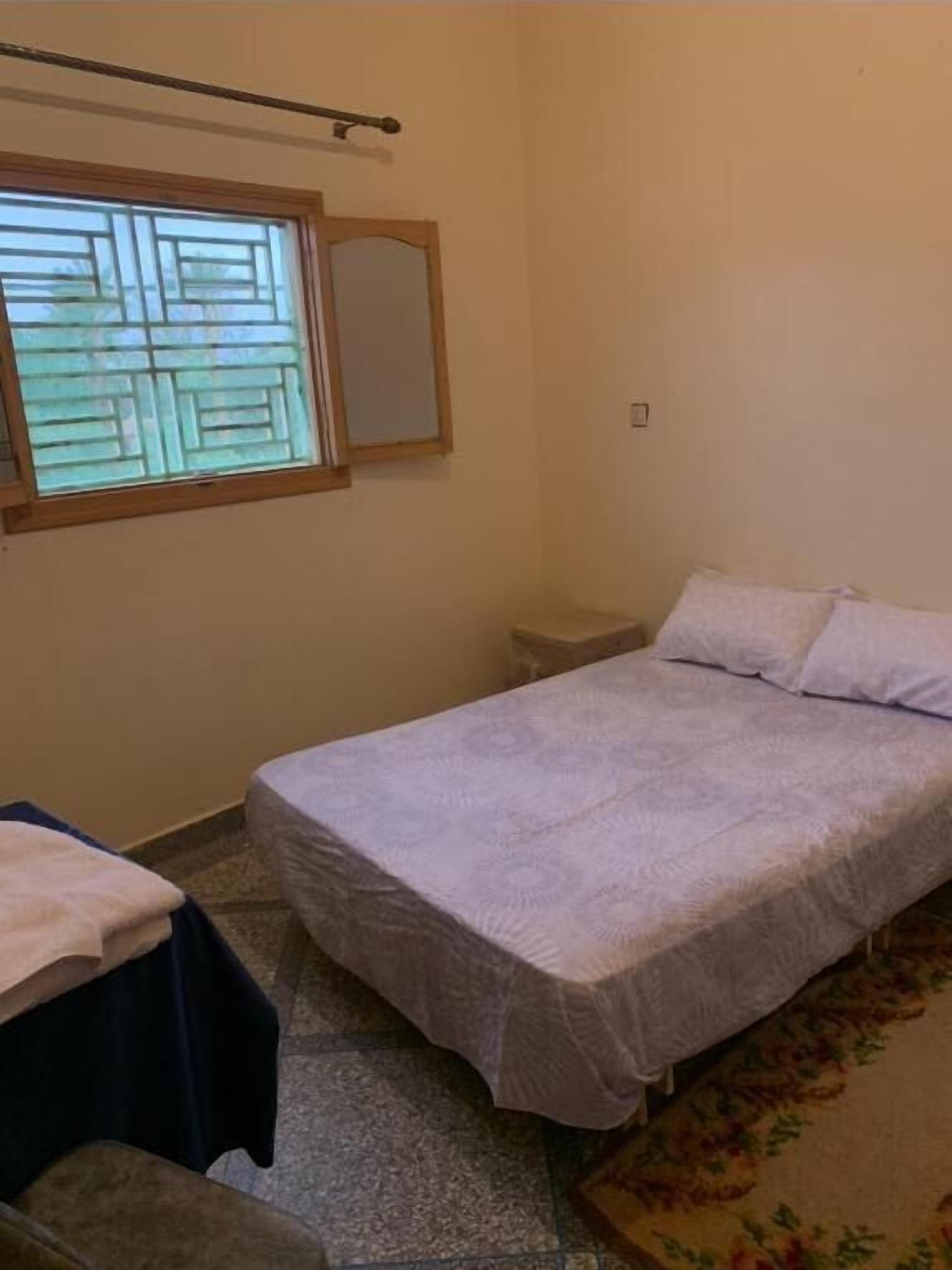 A bedroom with a bed, a nightstand, a window with blinds, and a mirror on the wall.