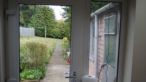 Uitzicht door een deurvenster op een tuinpad dat leidt naar een houten omheining en bomen.