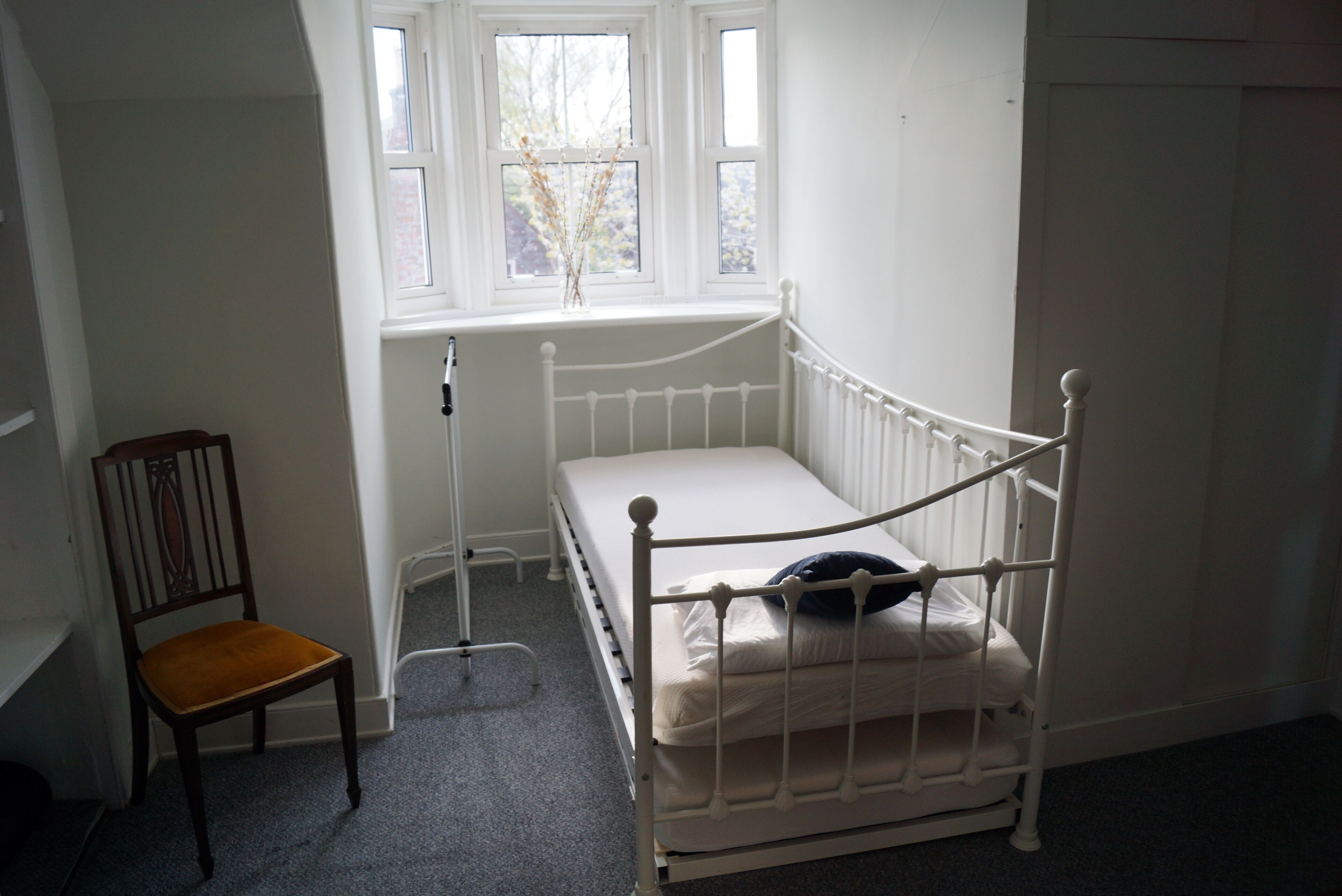A small, white, single-bed room with a window, a chair, and a radiator.