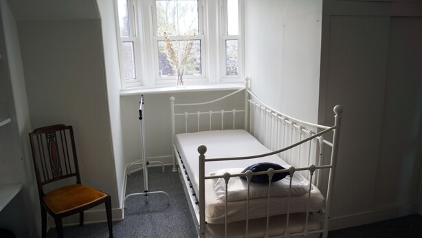 Een kleine, witte kamer met een enkel bed, een raam, een stoel en een radiator.