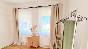 A room with a wooden floor, a small wooden side table with a globe and dried plants, a clothing rack with a green towel, and sheer curtains on a window.