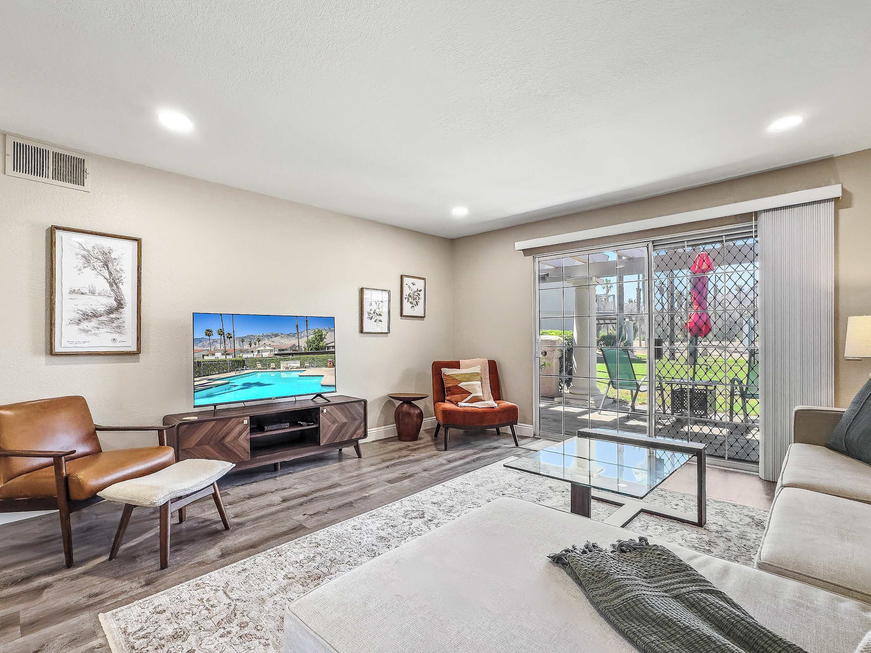 A living room with a flat-screen TV, a brown leather chair, a glass coffee table, and a large window with a view of a pool and greenery.