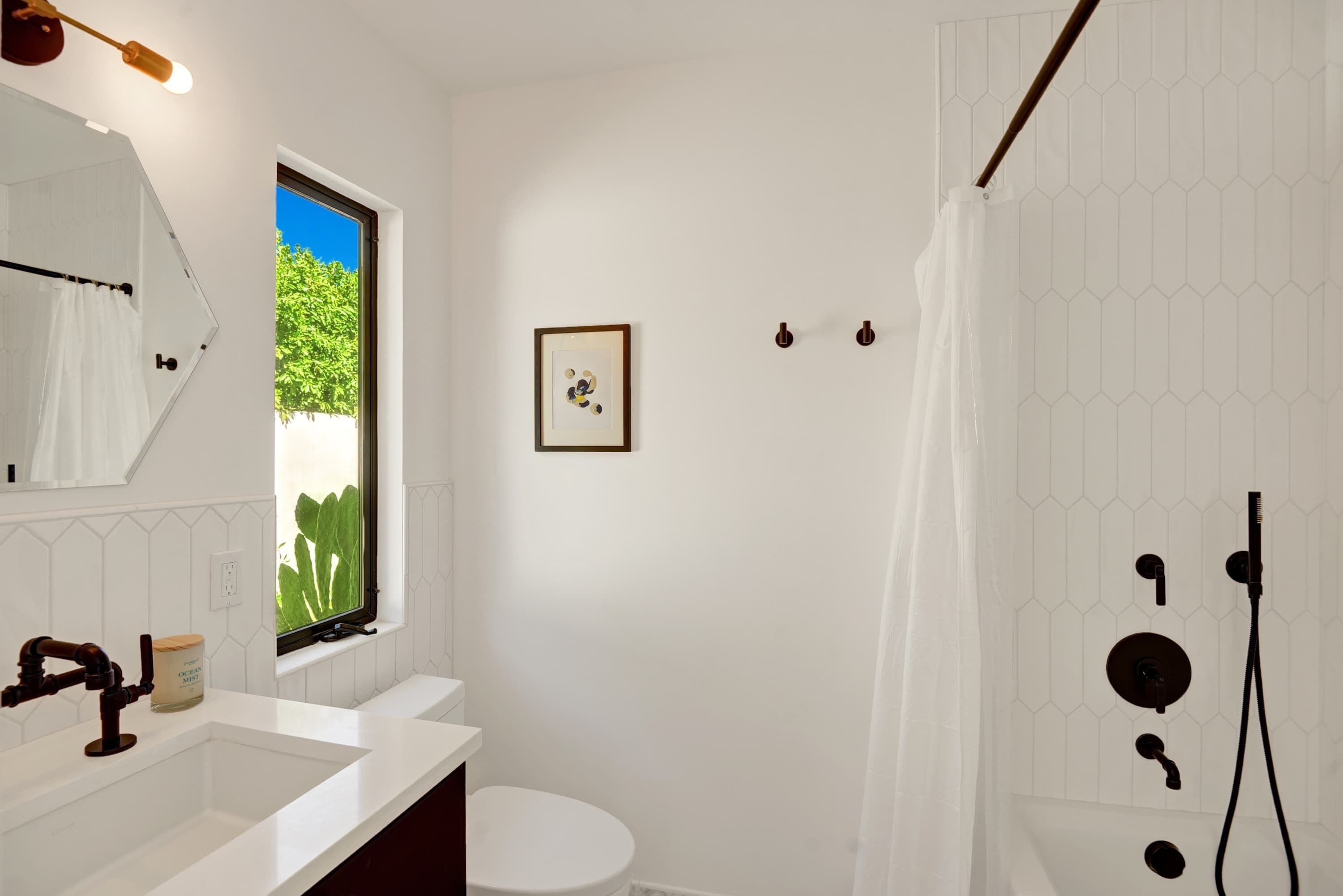 A bathroom with a white sink, toilet, and shower area, a window with a view of greenery, and a framed picture on the wall.