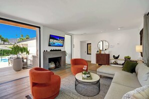 A modern living room with a fireplace, a television mounted on the wall, and a view of a pool and mountains. - Casa Palmera-Mid Century Modern (Palm Springs)