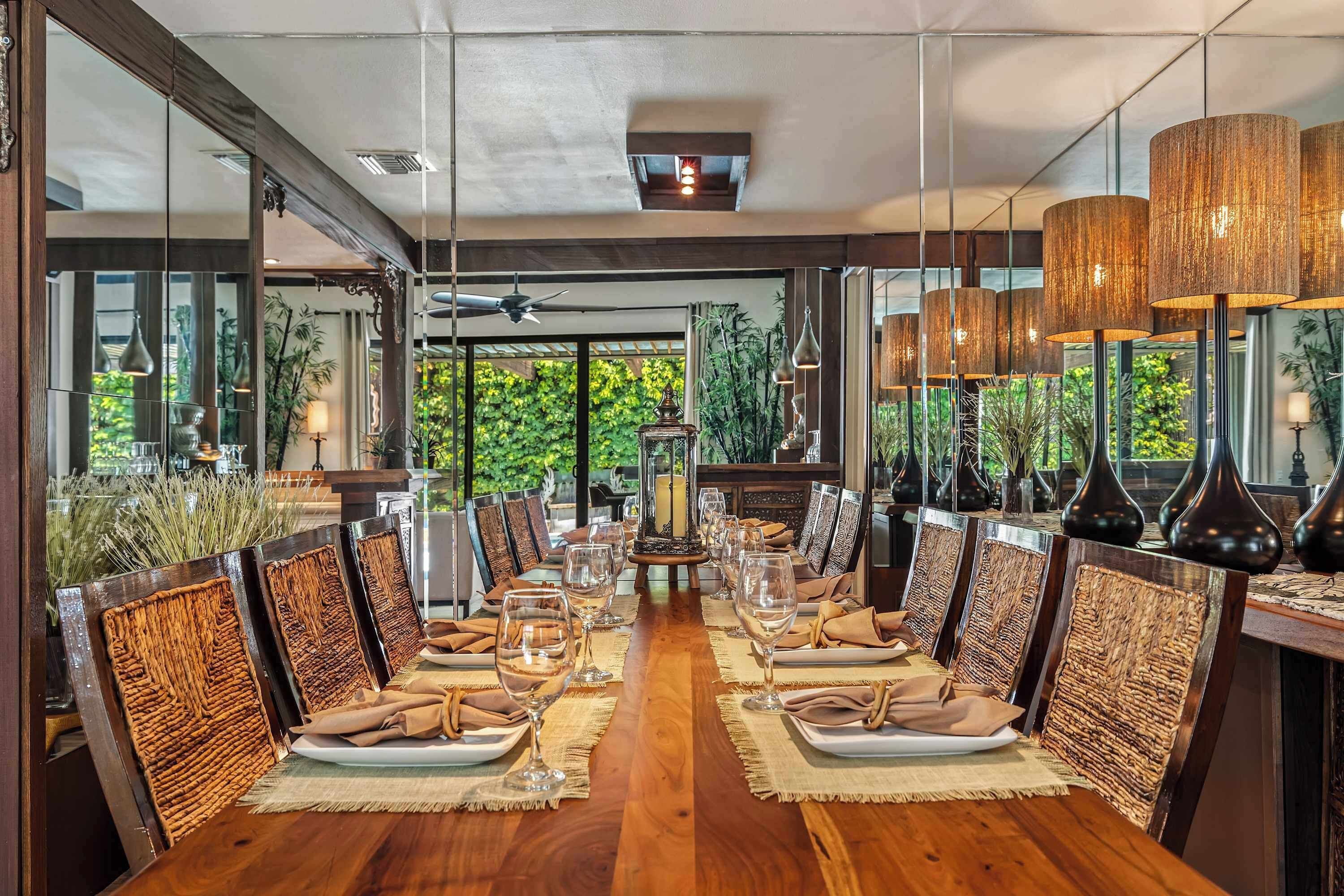 Comedor con una larga mesa de madera dispuesta para una comida, rodeada de sillas con respaldo de mimbre e iluminación colgante.