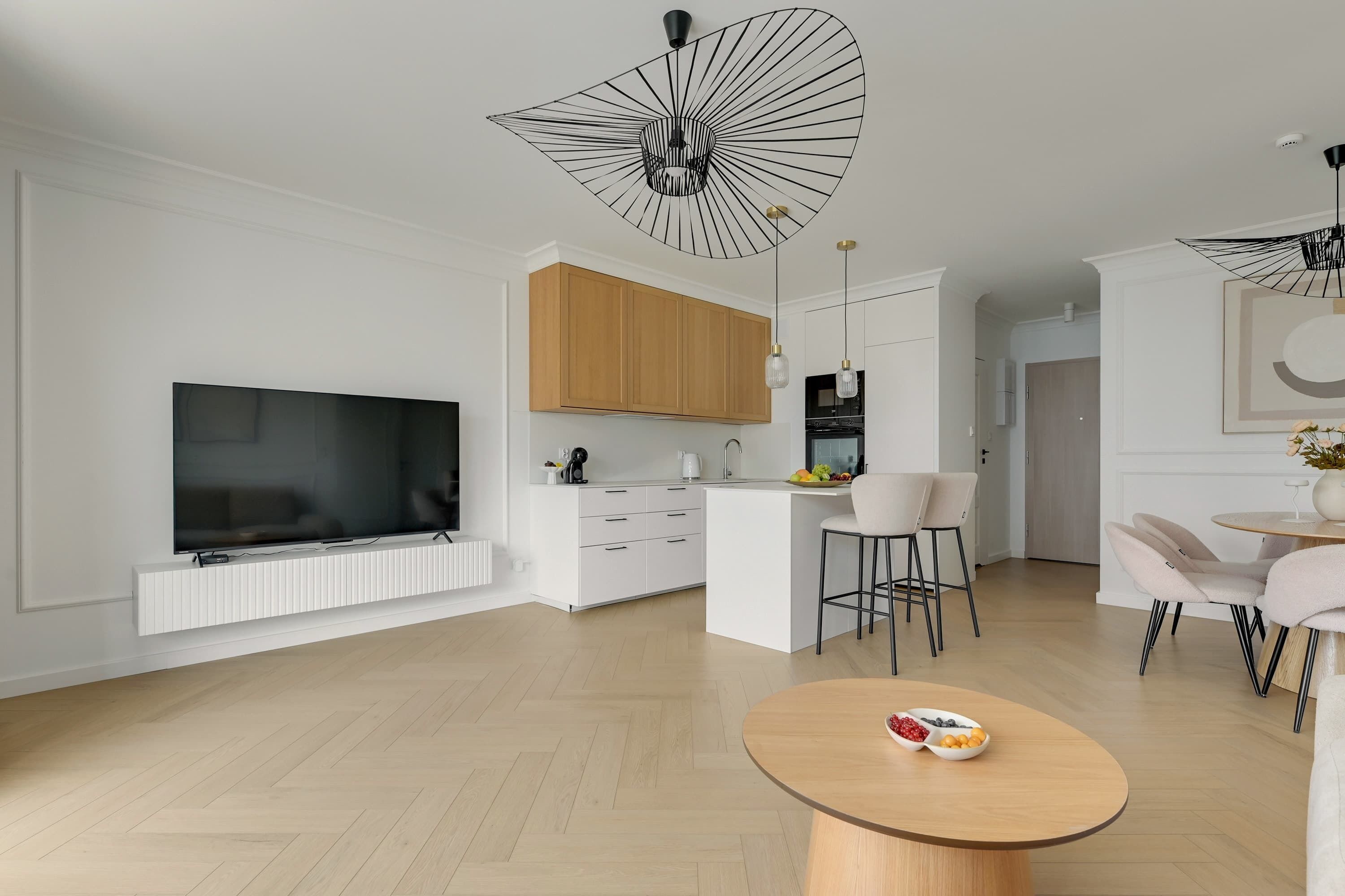A modern living room with a flat-screen TV, a dining area with a round table, and a kitchen with wooden cabinets.
