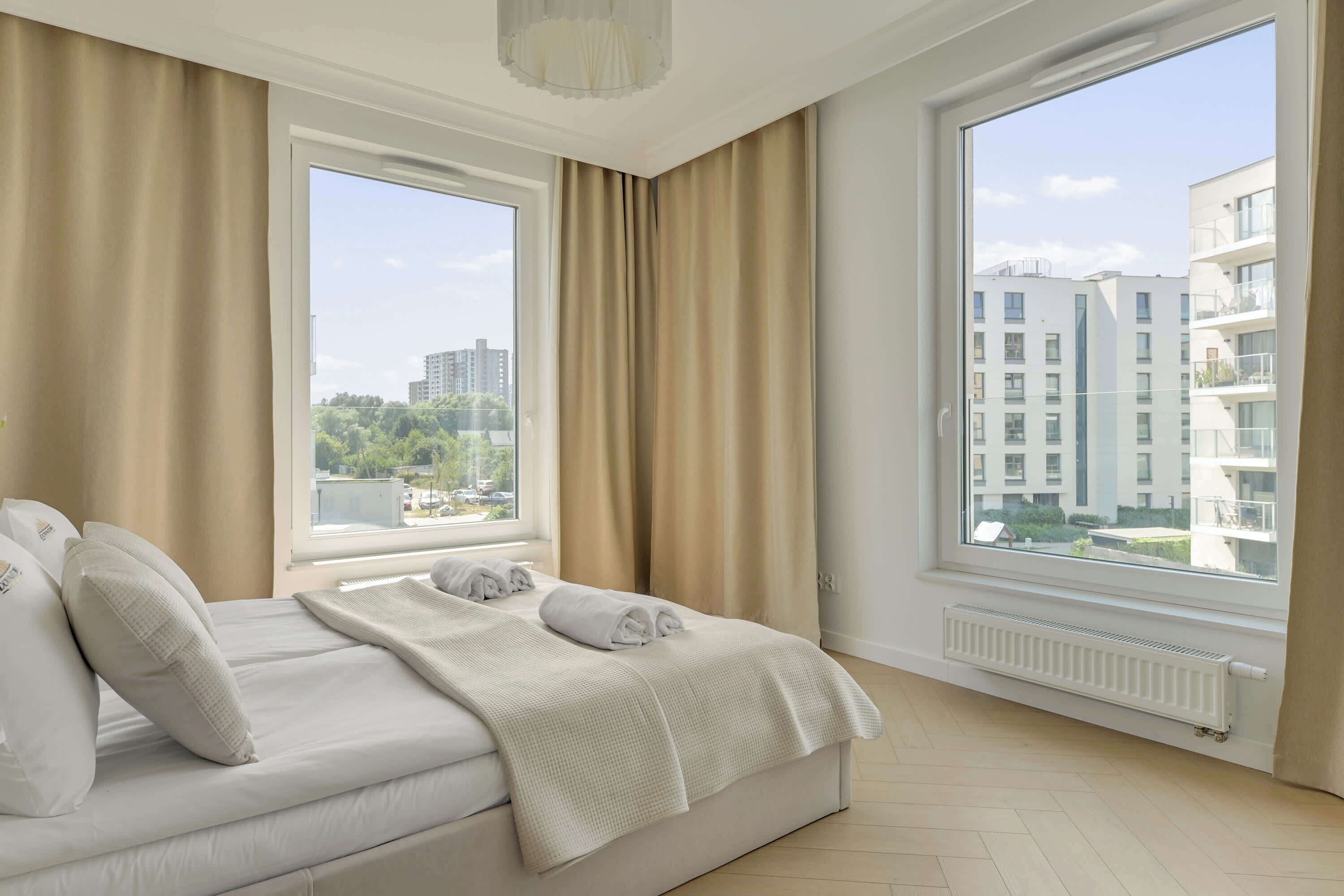 A hotel room with a bed, two windows, and a view of buildings.