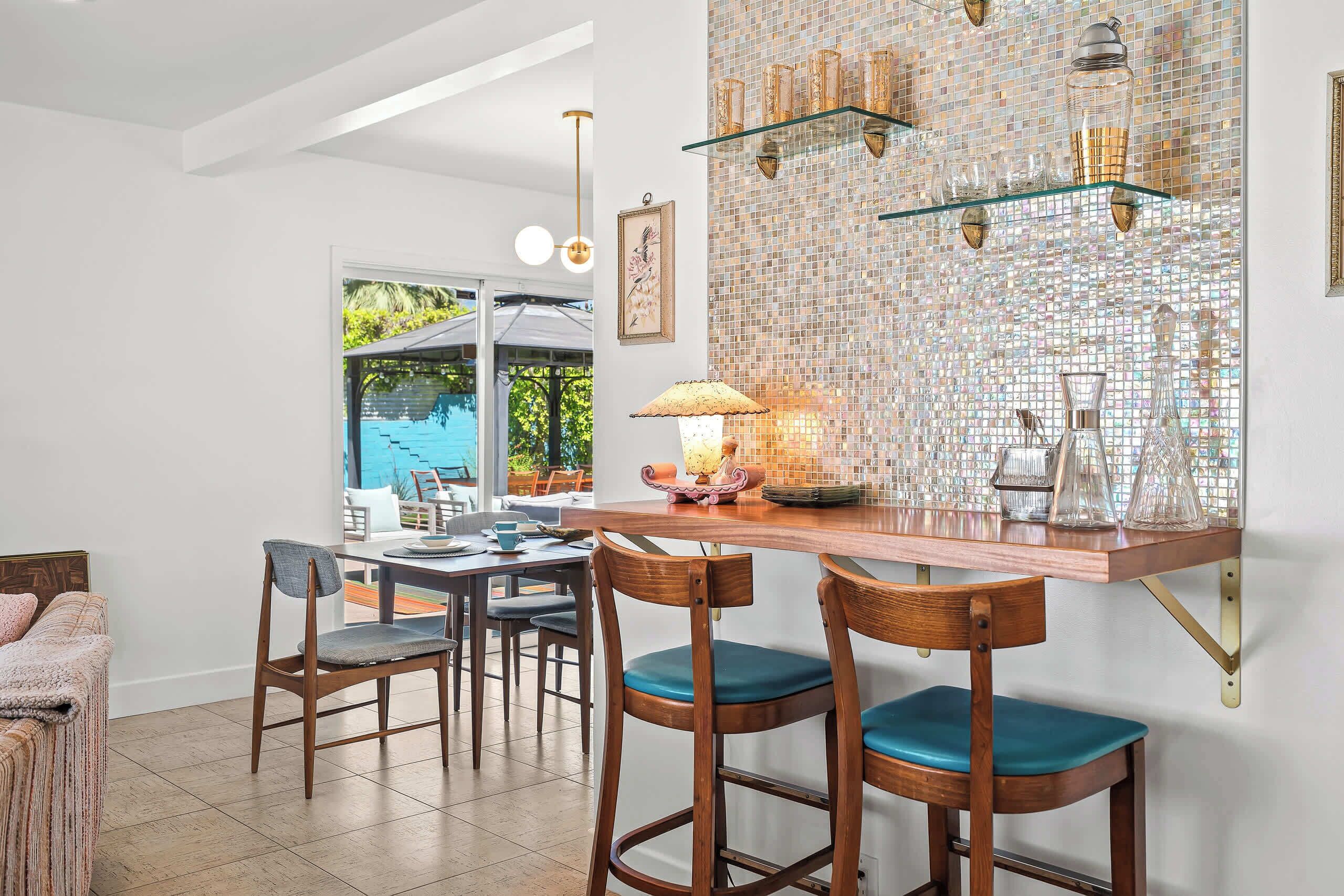 A dining area with a glass-top table, bar stools, and a decorative wall with mosaic tiles.