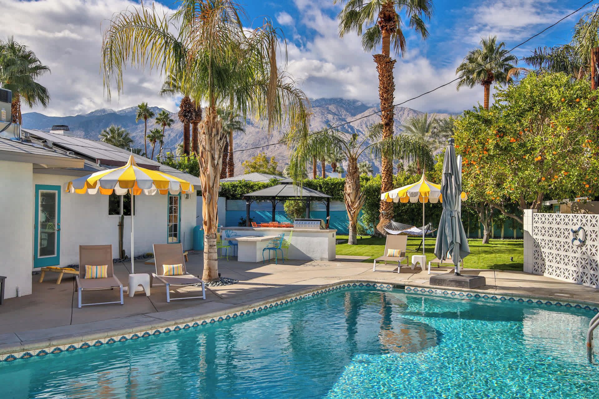 A swimming pool with lounge chairs and umbrellas, surrounded by palm trees and a mountain backdrop.