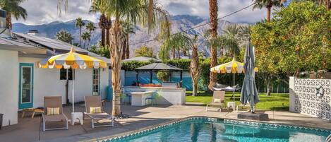 A swimming pool with lounge chairs and umbrellas, surrounded by palm trees and a mountain backdrop.