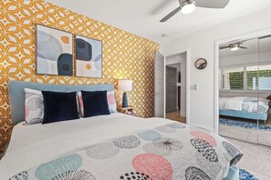 Ein Schlafzimmer mit strukturiertem Wandpapier, einem Bett mit weißer und gemusterter Bettdecke, einem Deckenventilator und Blick durch eine Glastür in einen anderen Raum.