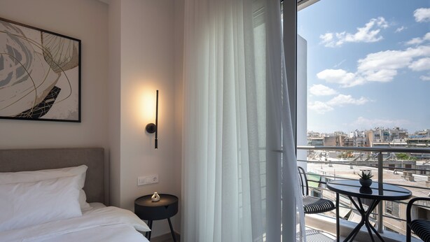 Habitación de hotel con cama, mesita de noche con un florero, luz de pared, cortinas translúcidas y un balcón con mesa y sillas.