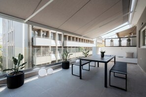 Un espacio al aire libre moderno con una mesa y bancos, plantas en macetas y vistas a un edificio con balcones.