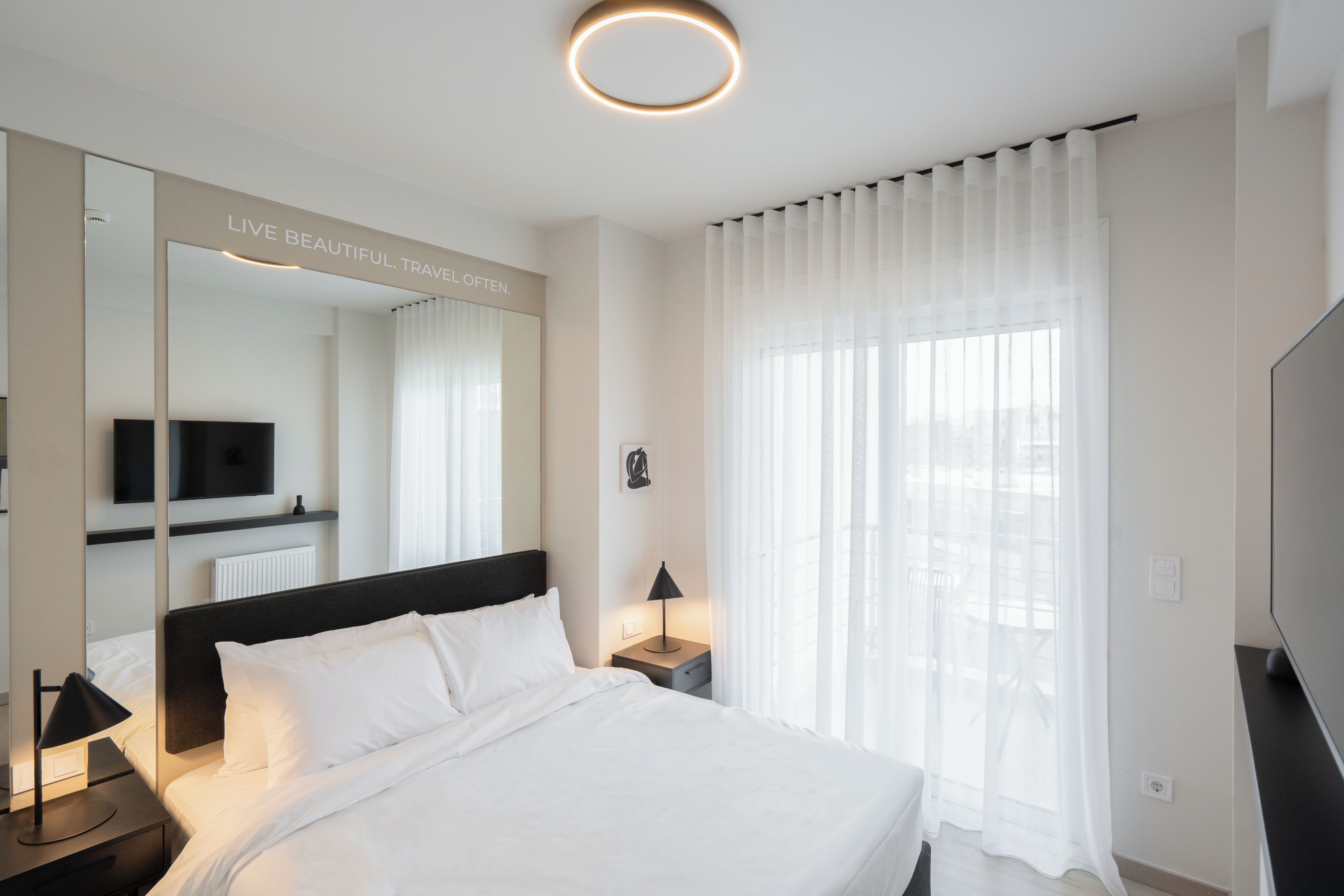 A hotel room with a bed, a nightstand, a lamp, a TV, a large mirror, and sheer curtains on a window.
