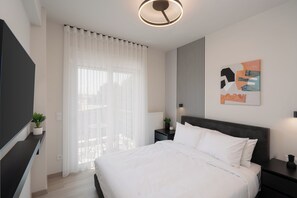 Un dormitorio moderno con una cama grande, un televisor de pantalla plana, una mesita de noche, una planta en maceta y una ventana con cortinas traslúcidas.