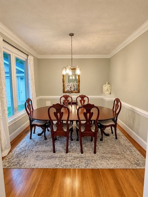 A dining room with a round table, six chairs, a chandelier, a mirror, and a vase with flowers.