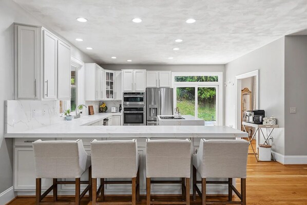 Una cocina moderna con armarios blancos, una isla grande con taburetes de bar, electrodomésticos integrados y vistas al exterior.
