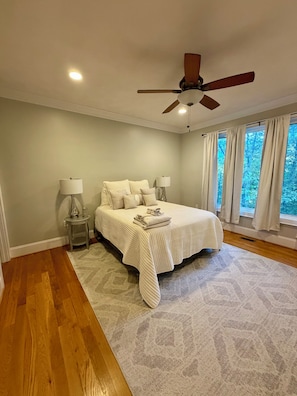 A bedroom with a bed, two bedside tables, a ceiling fan, and large windows with curtains.