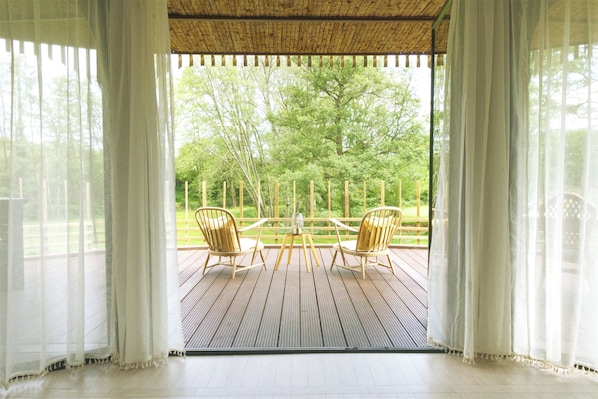 Une pièce dotée d’une terrasse en bois, de deux chaises, d’une table et offrant une vue sur un paysage verdoyant grâce à de grandes fenêtres.