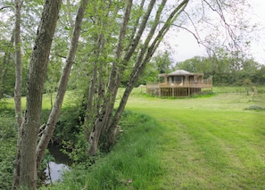Une cabane en bois dotée d’une terrasse, entourée d’arbres et bordée d’un champ d’herbe.