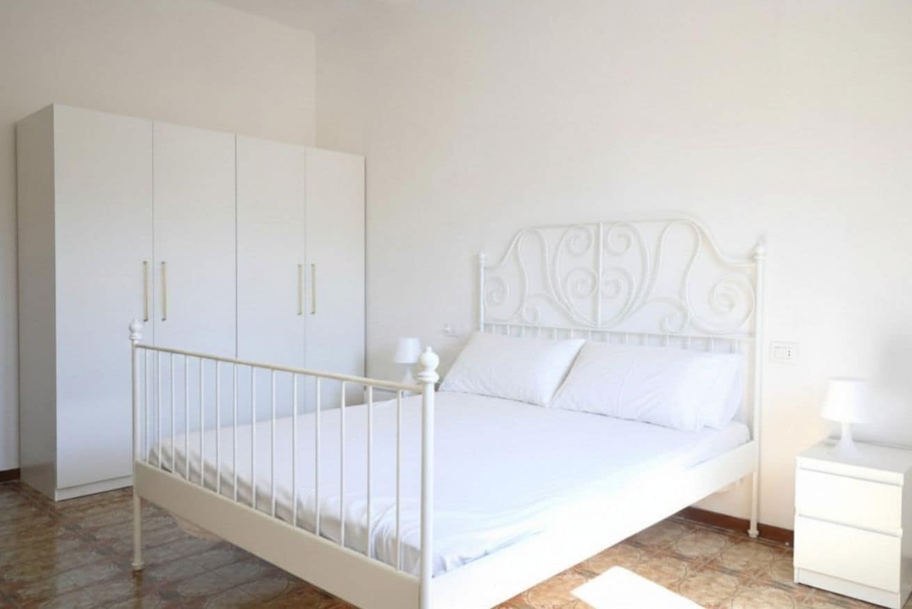 Uma cama de metal branca com cabeceira decorativa, um guarda-roupa branco e uma mesa de cabeceira com abajur.