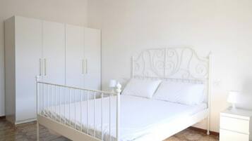 Uma cama de metal branca com cabeceira decorativa, um guarda-roupa branco e uma mesa de cabeceira com abajur.