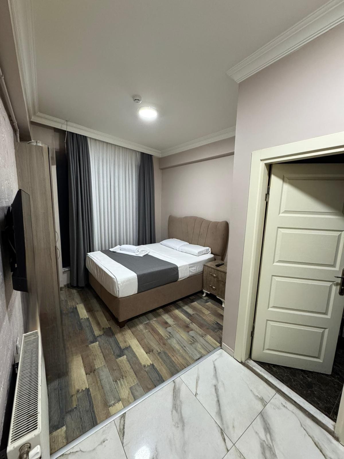 A hotel room with a bed, a bedside table, a TV, a radiator, and a door.