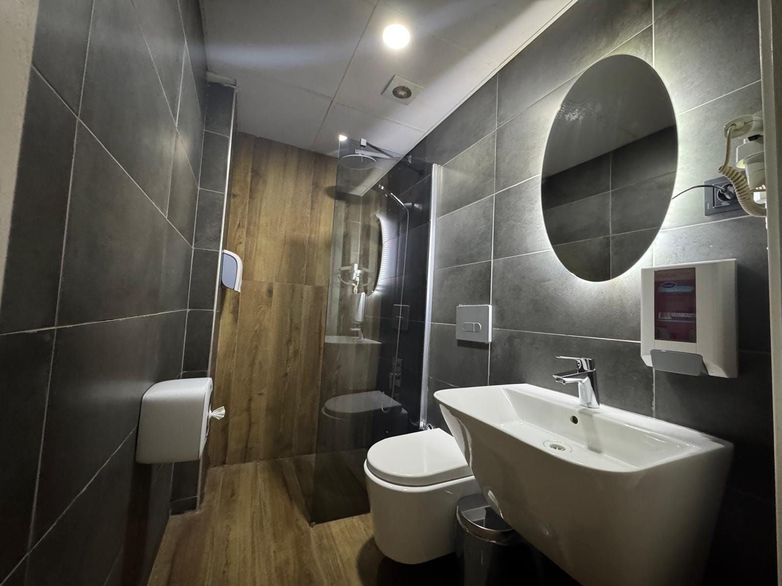 Un baño moderno con piso de madera, un lavamanos blanco, un inodoro y una zona de ducha.