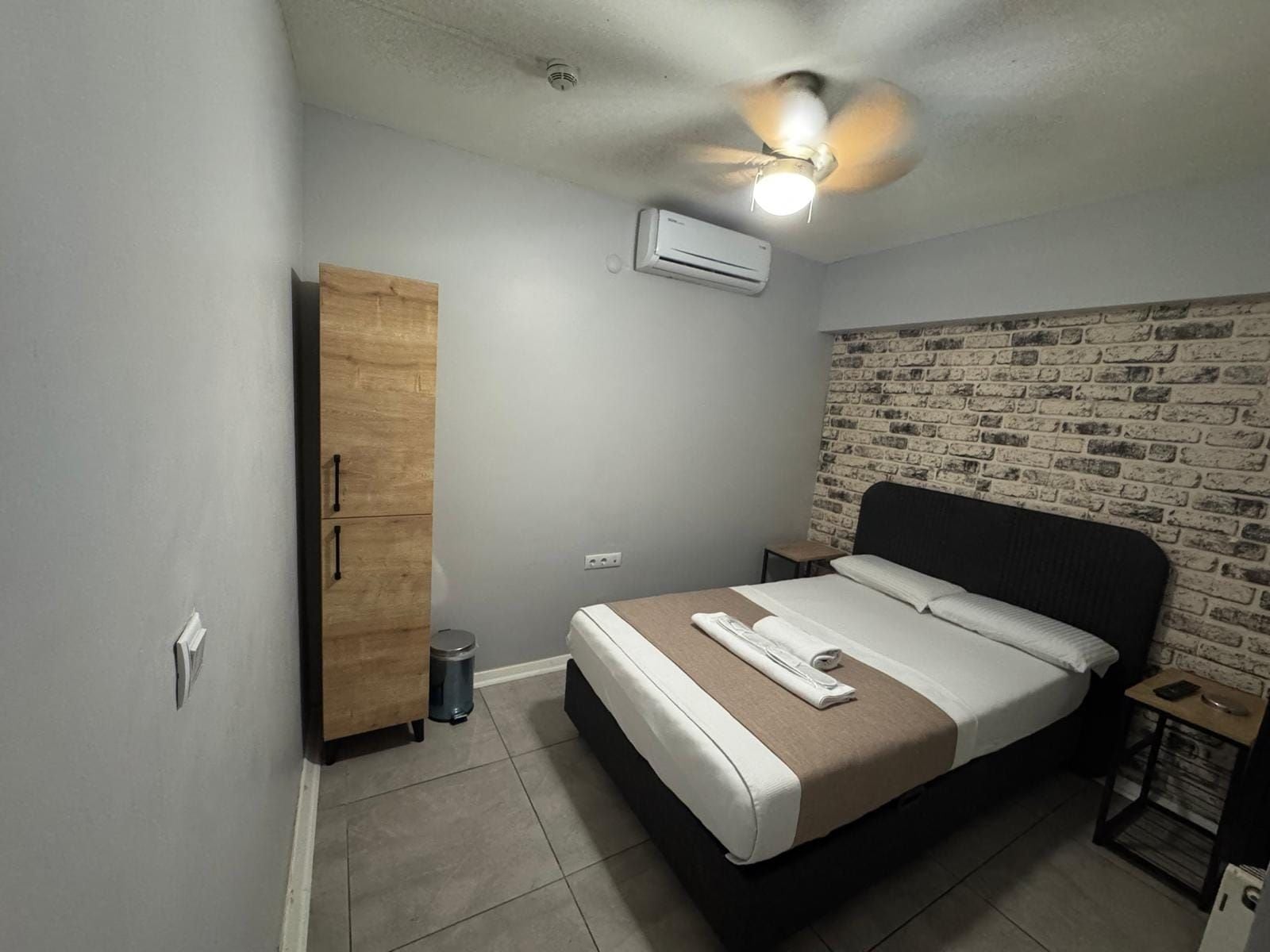 A bedroom with a bed, a wardrobe, a ceiling fan, an air conditioning unit, and a brick-patterned wall.