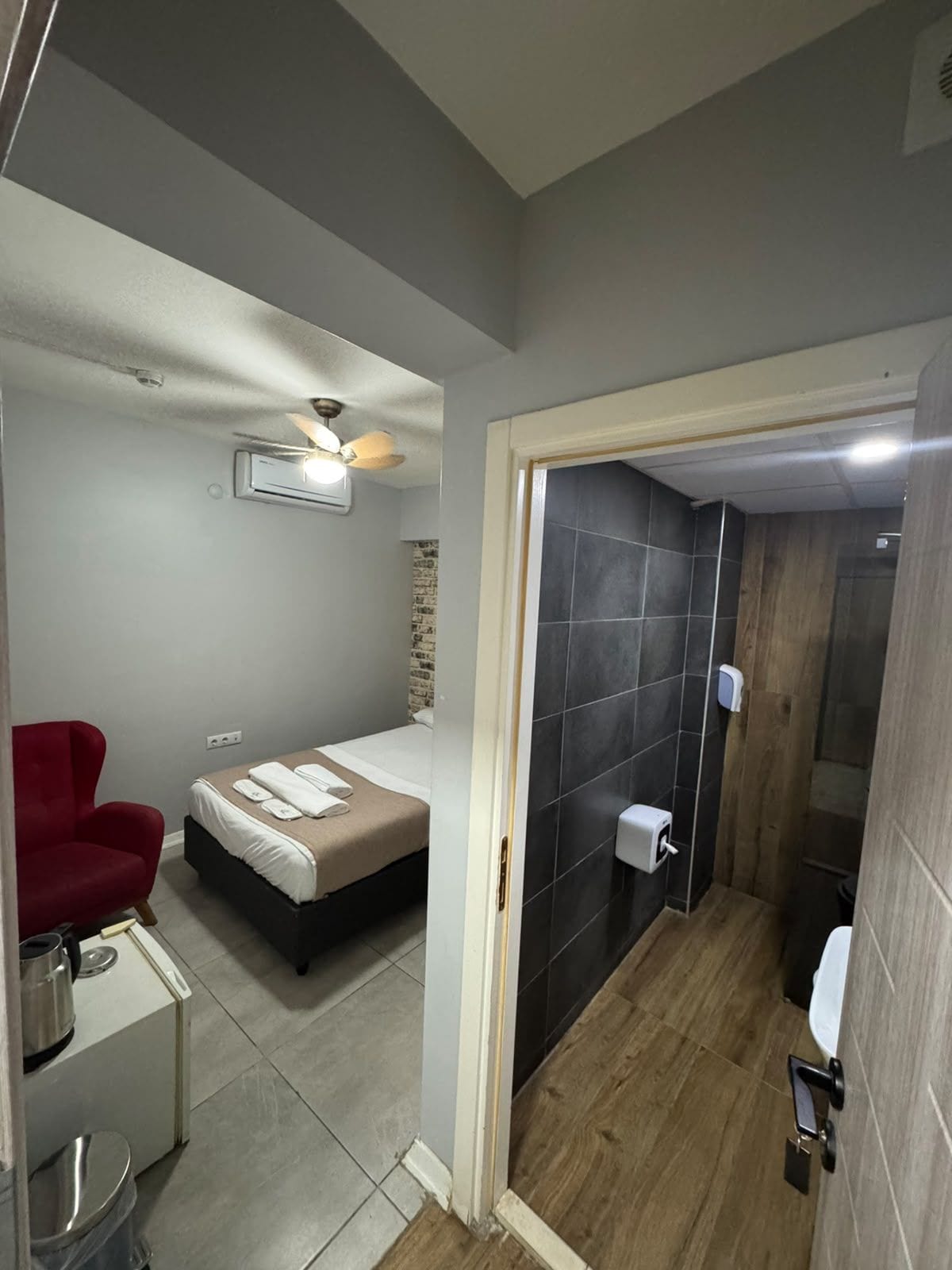 Habitación de hotel compacta con cama, una silla roja, una kitchenette pequeña y zona de ducha.