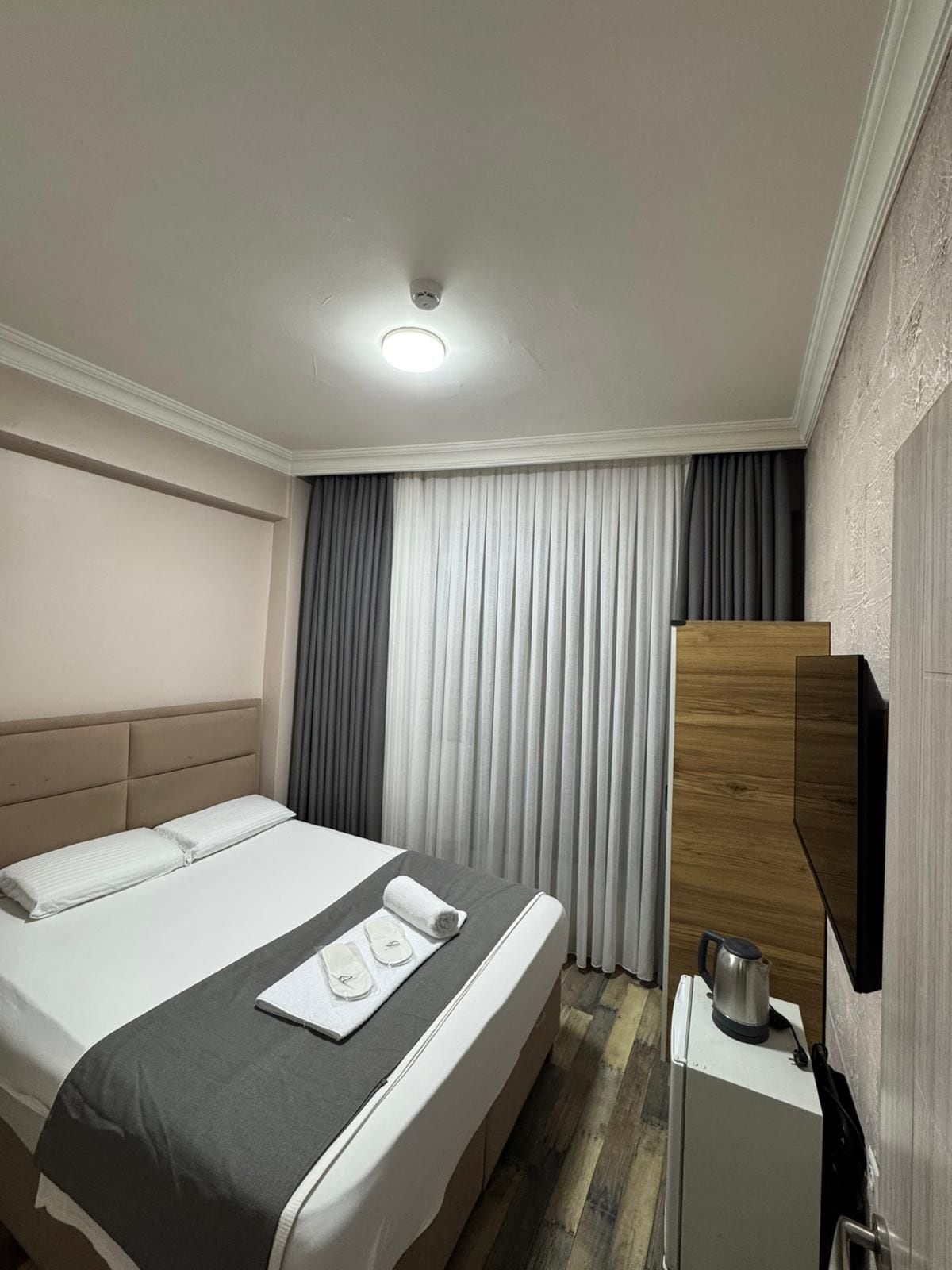 A hotel room with a bed, a bedside table with a kettle, and a large window with curtains.